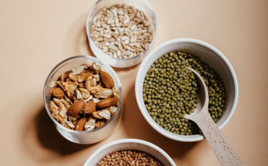 An overhead view of assorted nuts and grains in bowls with a spoon on a neutral background.