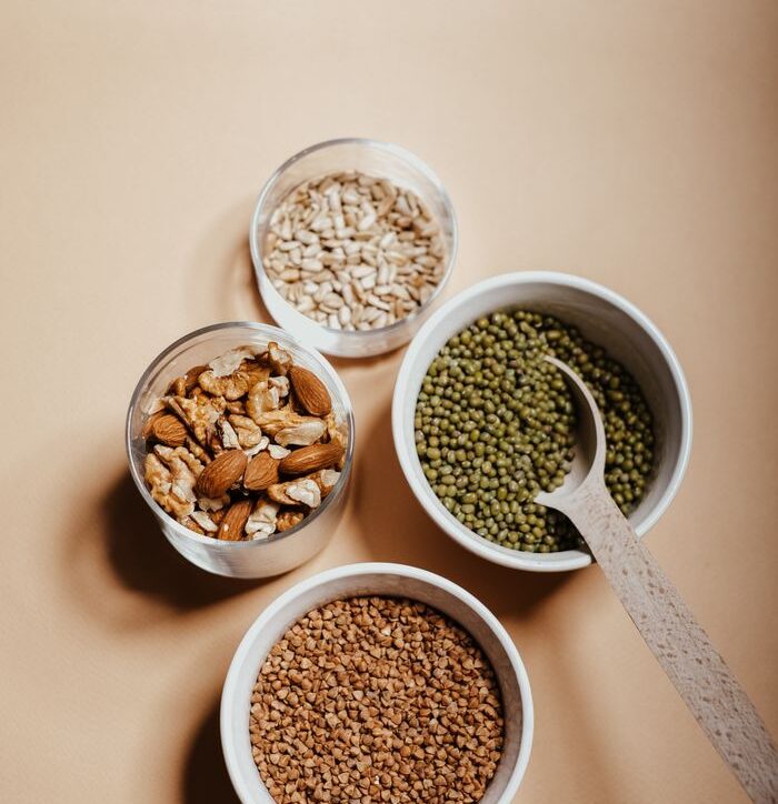 An overhead view of assorted nuts and grains in bowls with a spoon on a neutral background.